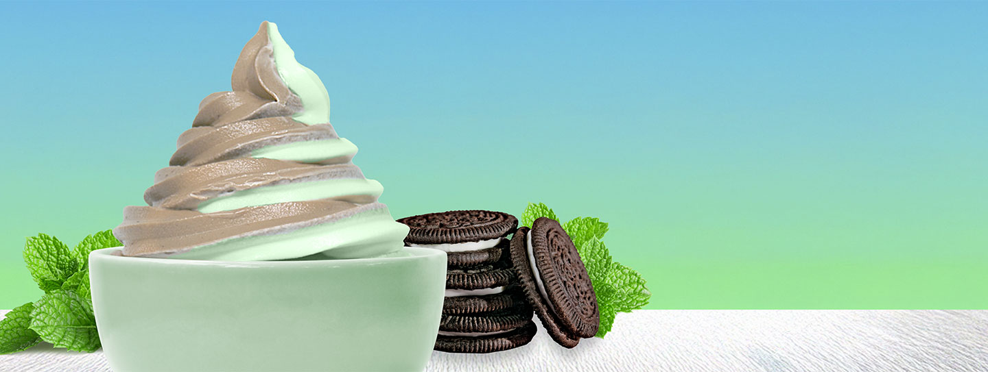 A cup of frozen yogurt alongside mint leaves and chocolate and cream cookies.