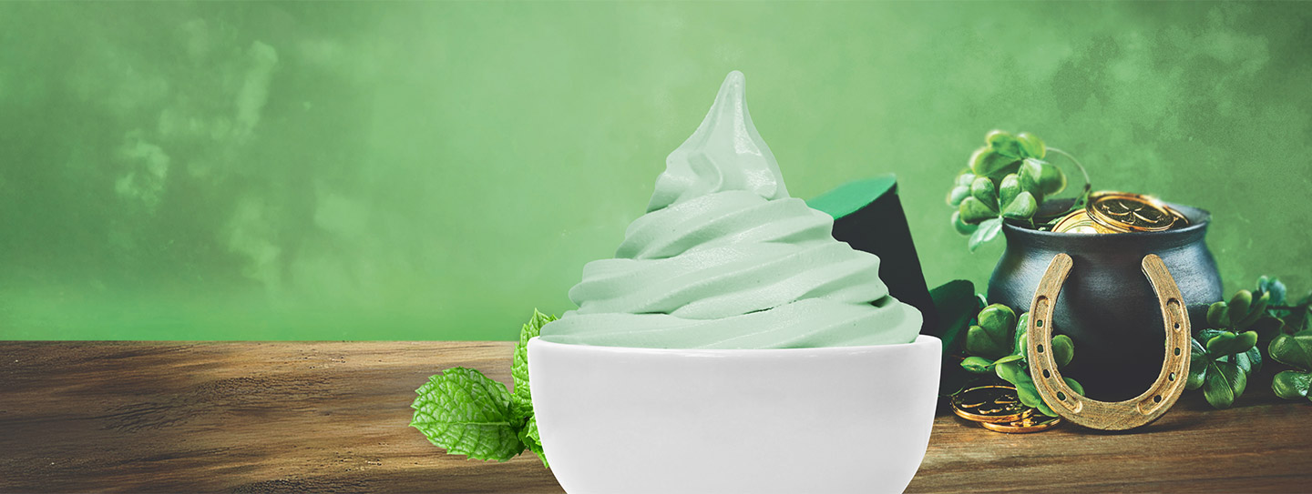 A cup of frozen yogurt alongside mint leaves, clovers and a pot of gold.